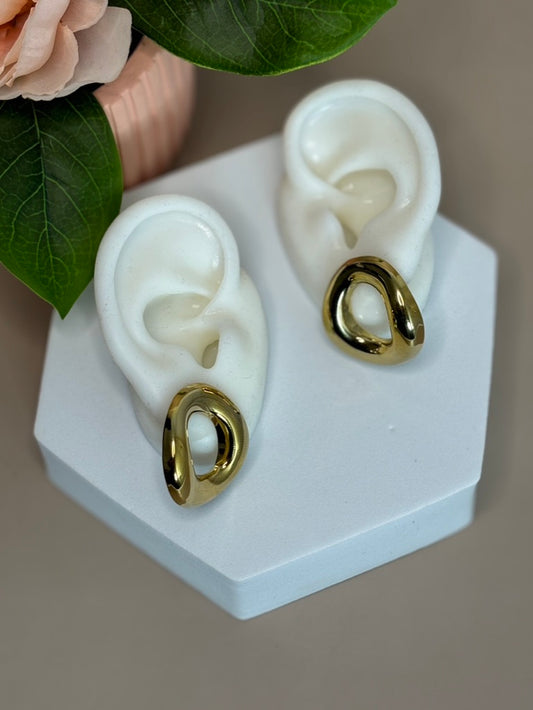 A pair of gold-colored stud earrings with a circle design displayed on a white stand, with a pink flower and green leaf in the background.
