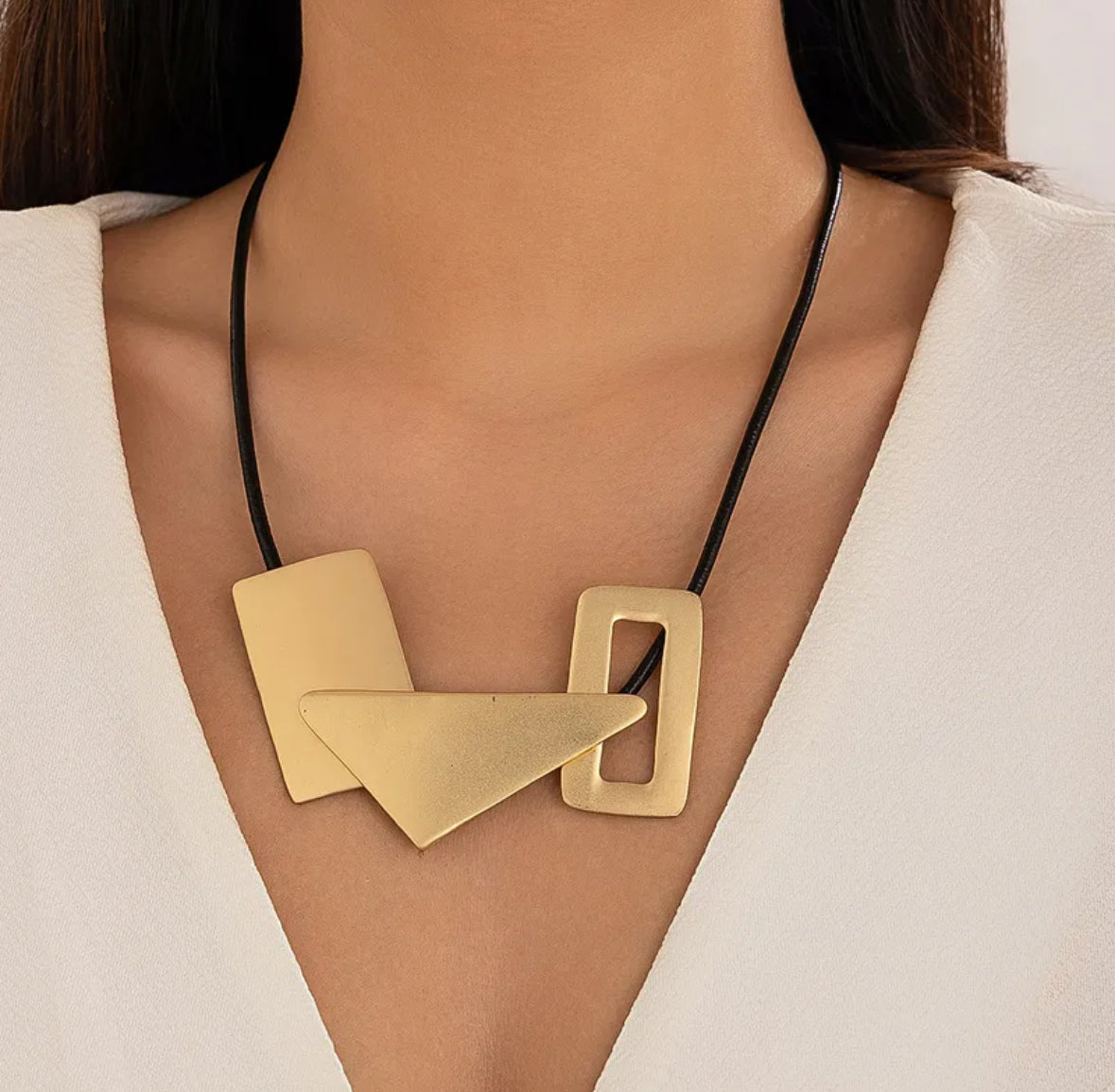 A close-up of a model wearing a modern geometric statement necklace. The necklace features three matte gold-toned metal shapes (a rectangle, a triangle, and an open square) connected by a thin black cord.