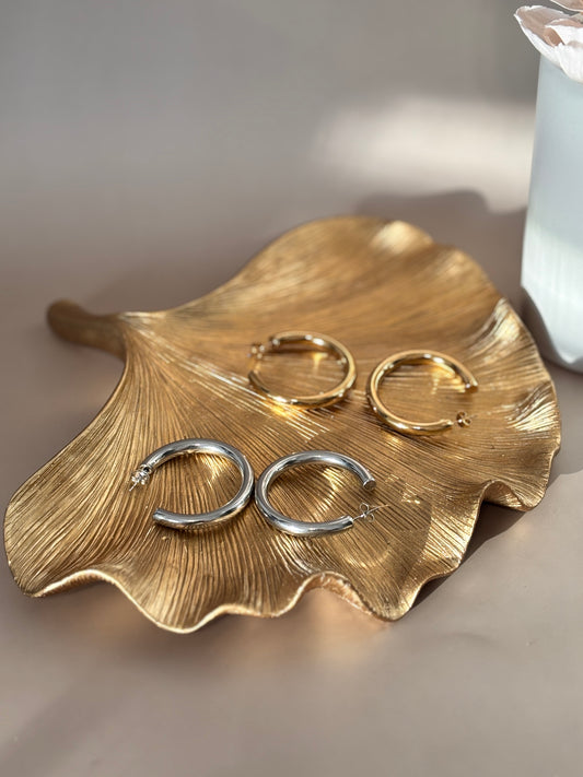 A pair of silver hoop earrings next to a pair of gold hoop earrings, placed on a gold-tone dish.