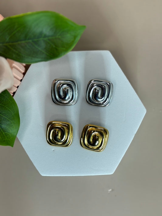 A pair of spiral charm stud earrings in gold and silver colors displayed on a white surface with a leaf in the background.