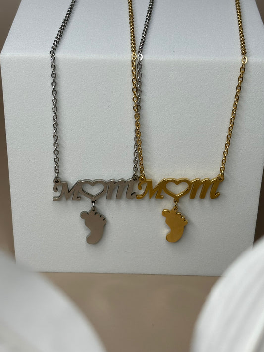 Two necklaces displayed on a pedestal, one in gold and the other in silver color, both featuring a stainless steel chain and a footprint design with the words 'Mom' and 'Love'.