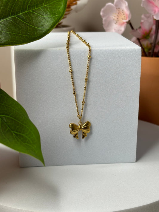 A gold-colored stainless steel necklace with a gold-plated bow-shaped pendant, displayed on a white background with green leaves in the background.