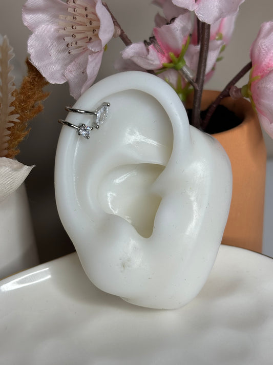 A silver-colored ear cuff with a double line design and crystals on the top, displayed on a surface with a background of flowers.