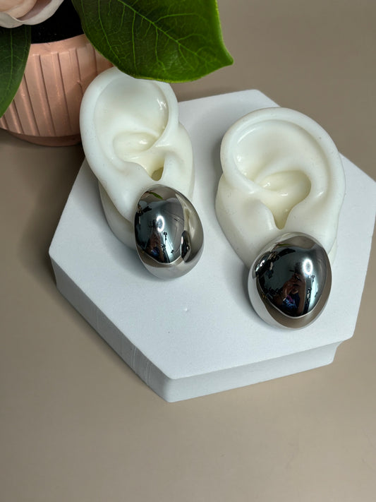 A pair of oval-shaped stainless steel earrings displayed on a white ear mold, with one earring reflecting light due to its metallic surface.