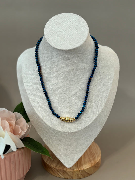 Blue color necklace displayed on a mannequin's neck, featuring artificial crystals with a mix of colors including gold and silver tones, with a background that includes a wooden frame and a pink floral decoration.