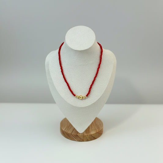 Red color necklace displayed on a mannequin's neck, featuring artificial crystals with a mix of colors including gold and silver tones, with a background that includes a wooden frame and a pink floral decoration.