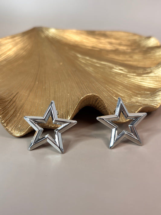 Two star-shaped earrings, one in gold and one in silver color, displayed on stands against a neutral background.